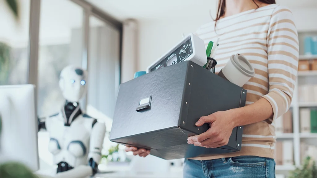 Robot at a computer while lady carries a box of her things leaving the office