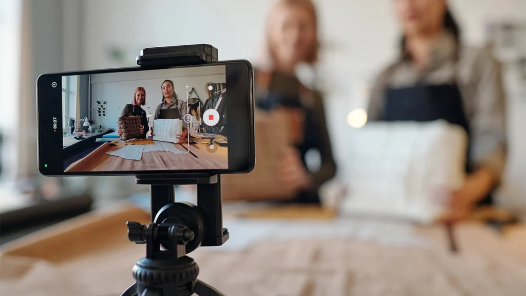 Two creative craftswomen on screen of smartphone standing on tripod in front of camera during masterclass on creating leatherwear