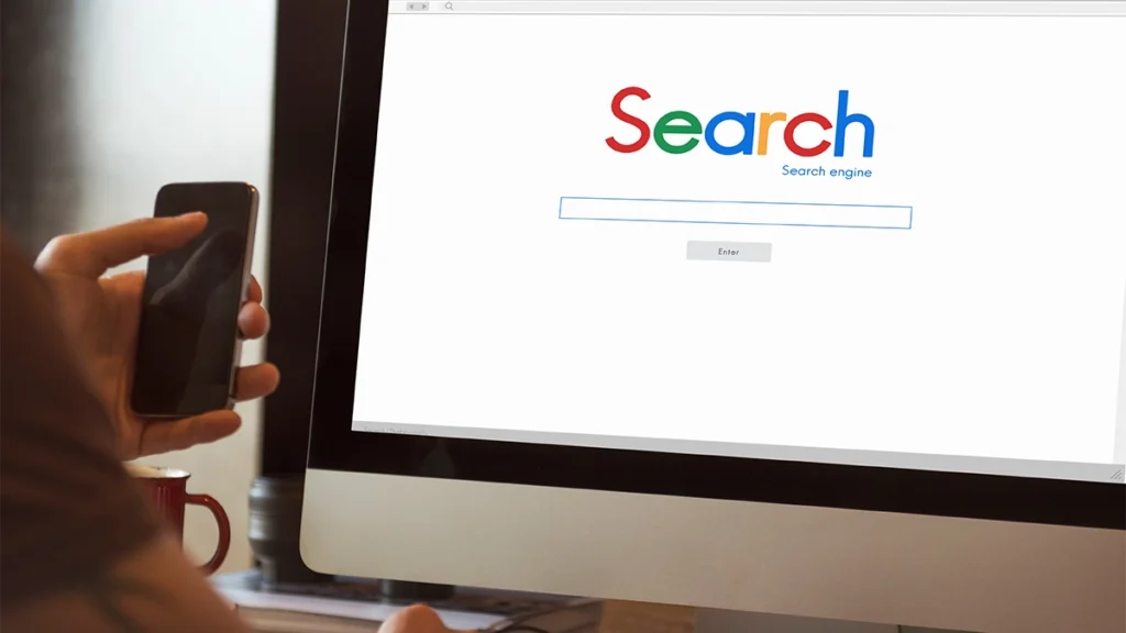 Man using search on a computer whilst holding a mobile phone