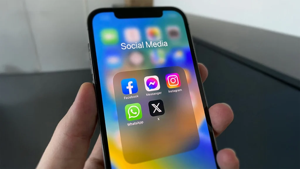 A hand holding a smartphone displaying a folder of social media apps including Facebook, Messenger, Instagram, WhatsApp and X.
