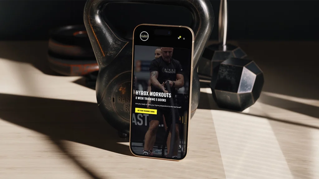 Phone with Hyrox Workouts website leaning against a kettlebell